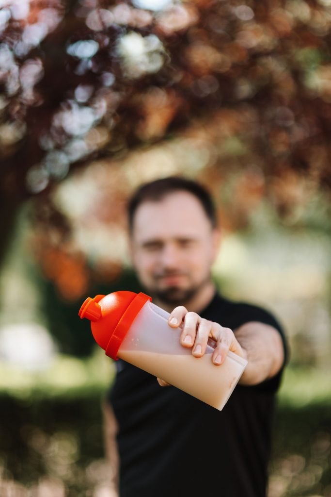 pexels-photo-4378522-4378522 Man holding out a protein shake bottle outdoors, wearing casual black shirt.